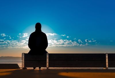 Silhouette man sitting on bench at beach against sky during sunrise