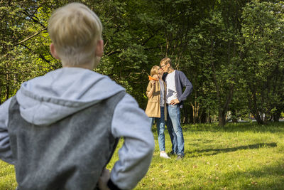 Back view of emotional caucasian boy looking at kissing parents in park. kid's jealousy. mom hugs
