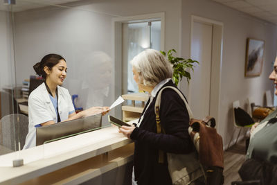 Smiling receptionist explaining form filling procedure to patient through transparent shield in clinic