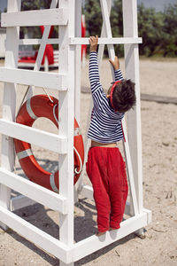 Child boy in striped clothes and red pants  standing on beach. white lifeguard tower, with a circle