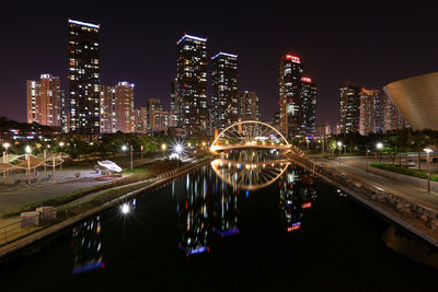 Illuminated modern buildings by river against sky at night