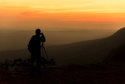 Silhouette man photographing on landscape against sky during sunset