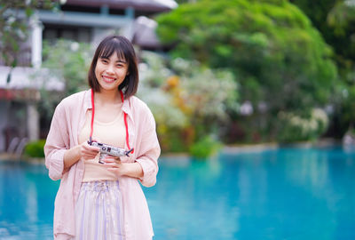 Woman holding camera while standing by swimming pool
