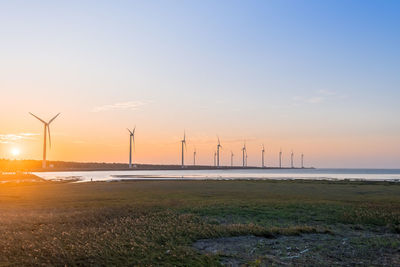 Wind turbines on field by sea against sky during sunset