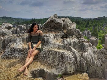 Young woman sitting on rock formations against cloudy sky