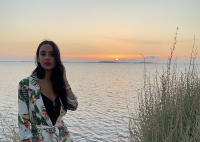 Young woman standing by sea against sky during sunset