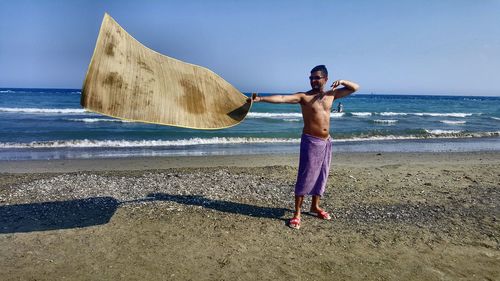 Full length of shirtless man holding mat while standing at beach against sky during sunny day