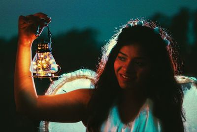 Close-up of smiling young woman holding illuminated string light while standing outdoors at dusk