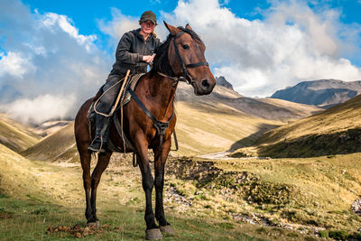 Side view of a shepherd rider on a horse standing on a mountain valley against sky with clouds