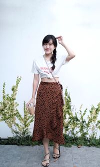 Woman standing against white wall wearing white tee-shirt  brown skirt holding ice coffee.