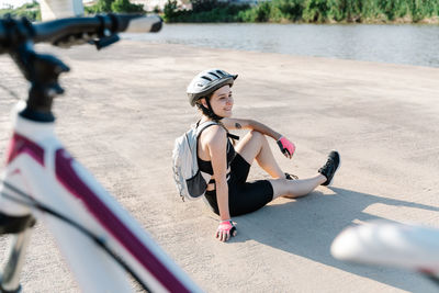 High angle side view of happy young woman in sportswear and helmet with backpack sitting on paved ground and resting after riding bike in summer day