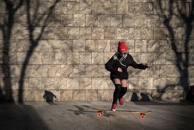 Full length of woman skateboarding on footpath against wall