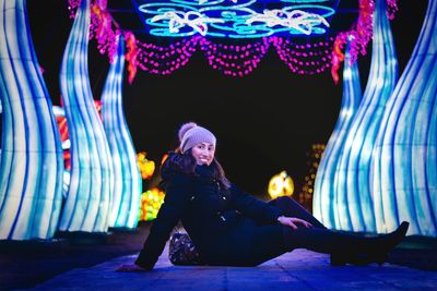 Portrait of smiling woman in warm clothing sitting on footpath amidst illuminated decoration
