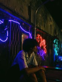 Man looking away while sitting against illuminated wall at night