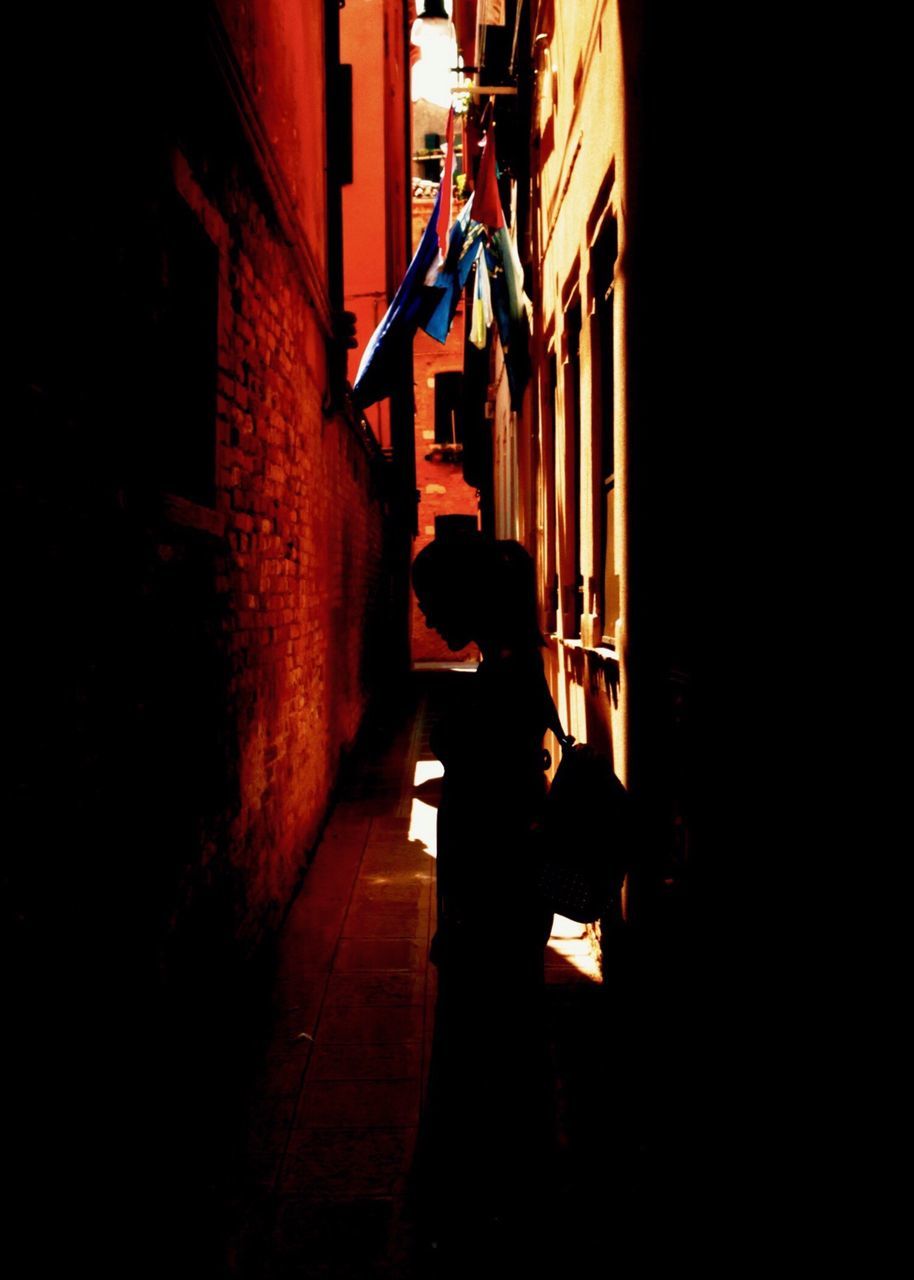 REAR VIEW OF WOMAN WALKING ON ALLEY AMIDST BUILDINGS AT NIGHT