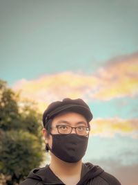 Portrait of young man in cap, eyeglasses and face mask standing against cloudy sunset sky