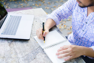 Midsection of man with book and pen by laptop on sidewalk cafe table