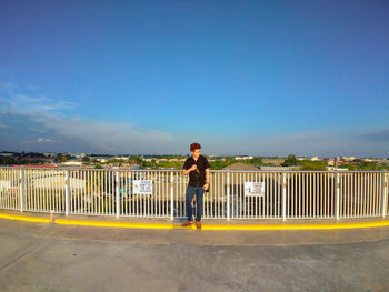 Full length portrait of man standing on railing against blue sky