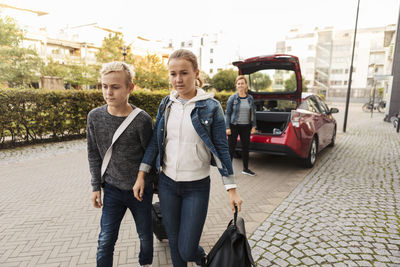 Siblings with suitcase walking while smiling mother standing by car on road