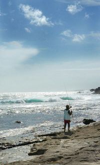 Rear view of man fishing at sea shore against sky