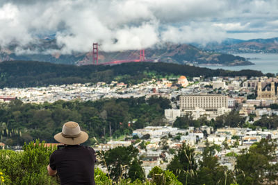 Rear view of man by cityscape and golden gate bridge against cloudy sky