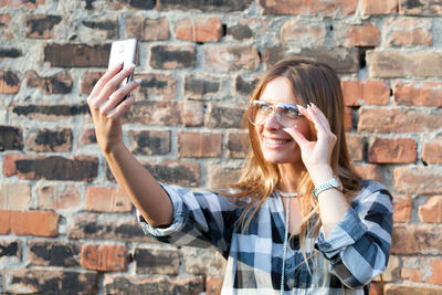 Mid adult woman taking selfie with smart phone while standing against brick wall