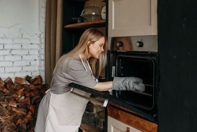 A young woman in an apron puts a baking tray in the oven in the kitchen at home