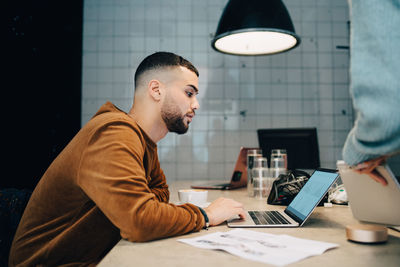 Side view of young male hacker using laptop at desk in small office