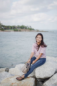 Portrait of smiling young woman sitting on rock against sea