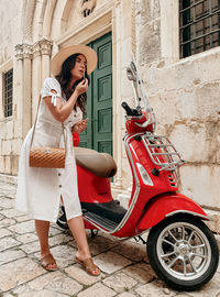 Beautiful young woman wearing white dress putting on lipstick while looking in mirror of red moped.