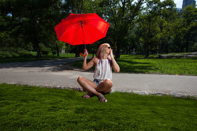 Full length of woman with red umbrella sitting at park