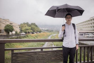 Portrait of man with umbrella standing on bridge during rainy season