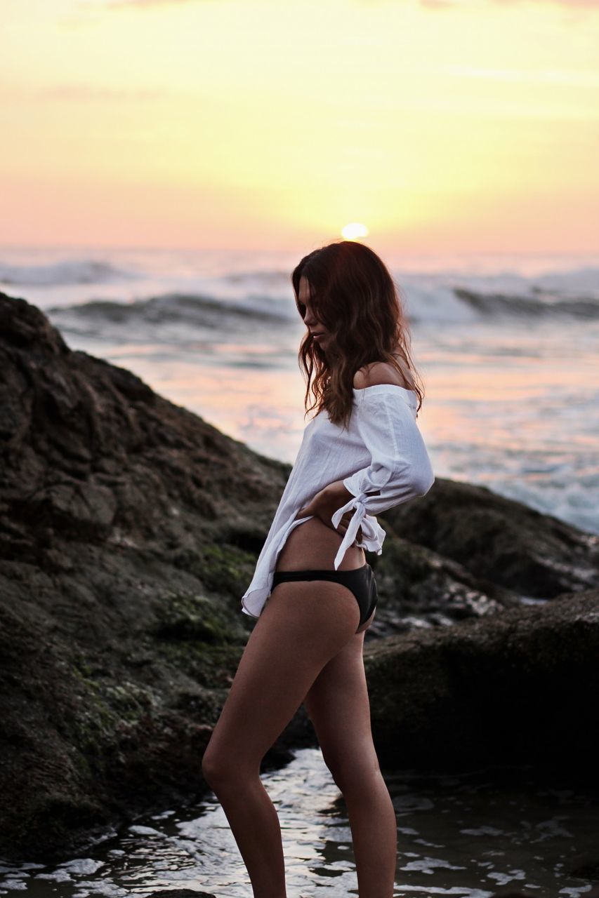 one person, water, sea, female, adult, sunset, women, sky, land, young adult, beach, nature, clothing, beauty in nature, rock, hairstyle, limb, long hair, human leg, ocean, coast, holiday, vacation, bikini, trip, photo shoot, fashion, lifestyles, leisure activity, relaxation, wave, full length, sunlight, standing, swimwear, side view, dusk, summer, person, tranquility, environment, shore, outdoors, scenics - nature, brown hair, sun, sports, motion, idyllic, looking, portrait, solitude, horizon, travel destinations, travel, cloud, human hair