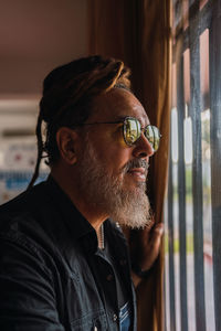 Side view of bearded mature male in casual outfit with sunglasses and dreadlocks standing near window while looking away thoughtfully in light room