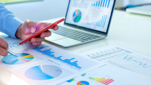 Cropped image of businessman using phone while analyzing graphs on table