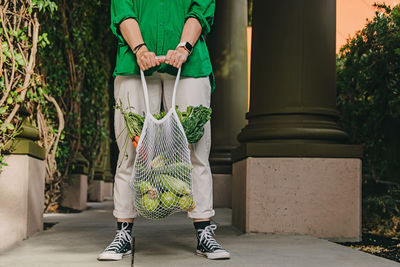Unrecognizable woman in green shirt holding a mesh bag with veggies on the street, close-up shot