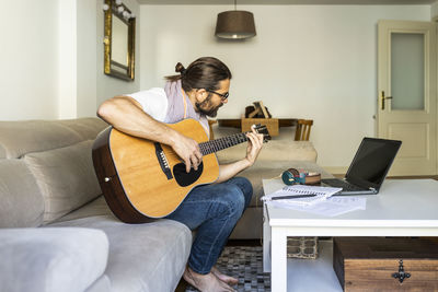 Side view of bearded creative male musician in casual wear sitting on sofa and playing acoustic guitar in modern living room