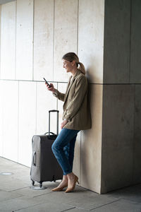 Smiling businesswoman using smart phone by luggage leaning on wall in tunnel