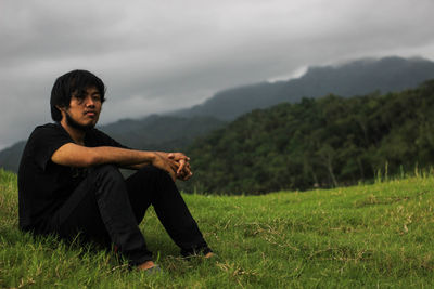 Young man sitting on field against sky