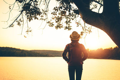Rear view of man standing by lake against sky during sunset