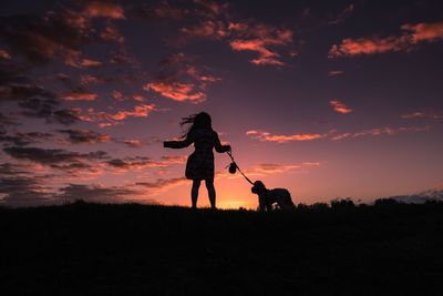 Rear view of girl with dog standing on silhouette field against sky during sunset