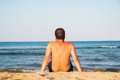 Rear view of shirtless man sitting at beach against clear sky