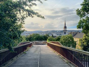 Empty walkway towards buildings and trees against sky