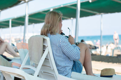 Young woman sitting on sun lounger on beach,putting on sunglasses and looking dreamily at the sea