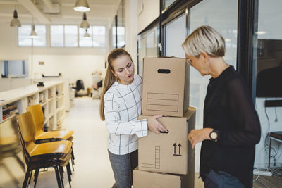 Mature businesswoman looking at female colleague carrying cardboard boxes in new office