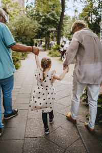 Grandparents holding hands of granddaughter while walking on footpath at street