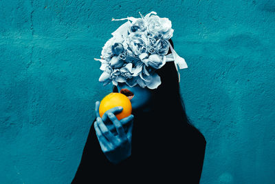Close-up of woman wearing flowers holding orange against wall