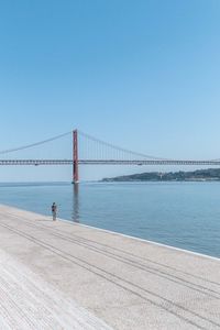 Man walking beside an ocean bay with a bridge in the background.