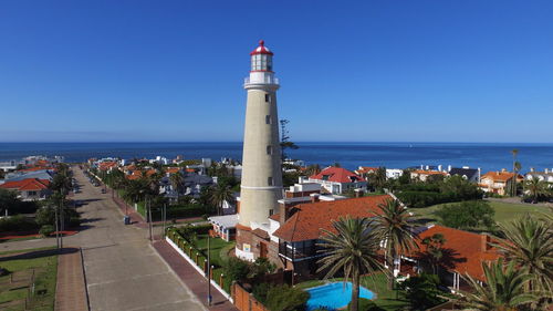 Lighthouse by sea against buildings against clear blue sky