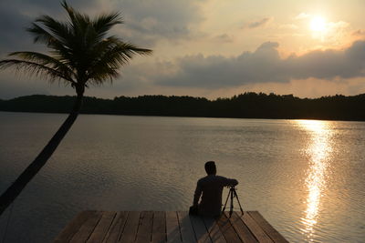 Rear view of silhouette man sitting by lake against sunset sky
